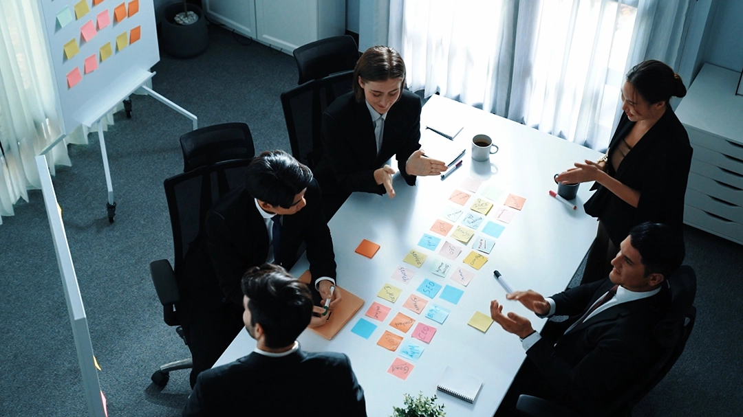 A group of professionals in suits discusses ideas over a table covered with colorful sticky notes in a modern office setting.